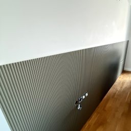 MZK MEBLE YURII MUZYKA - Montaż panelu dekoracyjnego z pionowymi żłobieniami na ścianie, widoczne gniazdka elektryczne w trakcie instalacji, parkiet na podłodze.