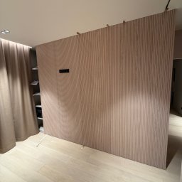 MZK MEBLE YURII MUZYKA - Drewniana ścianka działowa z pionowych paneli w jasnym odcieniu drewna, oddzielająca przestrzeń z widoczną półką i zasłoną, oświetlona punktowo, z widocznymi elementami montażowymi u góry.