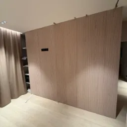 Drewniana ścianka działowa z pionowych paneli w jasnym odcieniu drewna, oddzielająca przestrzeń z widoczną półką i zasłoną, oświetlona punktowo, z widocznymi elementami montażowymi u góry.