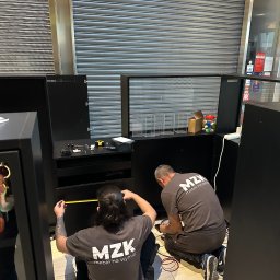 MZK MEBLE YURII MUZYKA - Dwóch monterów w szarych koszulkach z logo 'MZK meble na wymiar' mierzy czarne moduły meblowe w lokalu z zamkniętą metalową roletą.