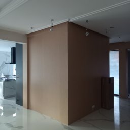 MZK MEBLE YURII MUZYKA - Wnętrze w trakcie wykańczania: widoczna ściana w kolorze beżowym, błyszcząca podłoga z marmurowym wzorem, fragment kuchni z szarymi szafkami i białym blatem, oraz kartony stojące przy oknie.