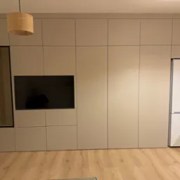 Beżowa zabudowa meblowa w salonie z wbudowanym telewizorem, przeszkloną witryną i lodówką, oświetlona wiszącymi lampami z abażurami z plecionki.