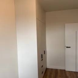 Wysoka, biała szafa wnękowa z czarnymi, minimalistycznymi uchwytami, dopasowana do sufitu, na tle białej ściany i drewnianej podłogi, obok białe drzwi z czarnym akcentem.