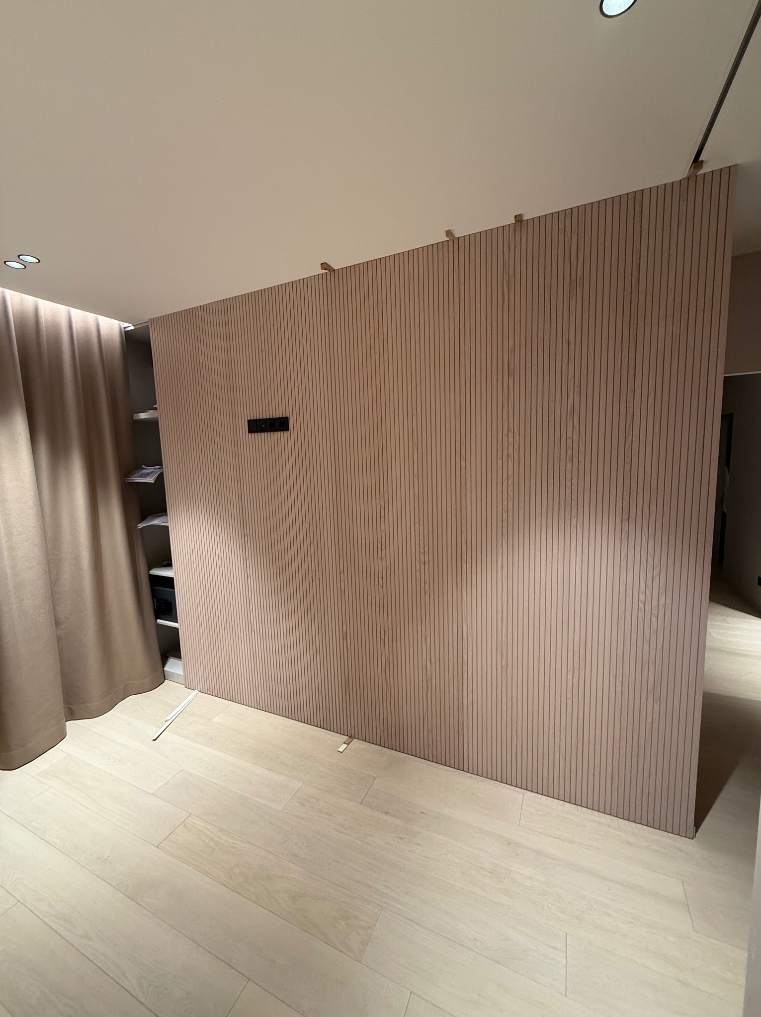 Drewniana ścianka działowa z pionowych paneli w jasnym odcieniu drewna, oddzielająca przestrzeń z widoczną półką i zasłoną, oświetlona punktowo, z widocznymi elementami montażowymi u góry.