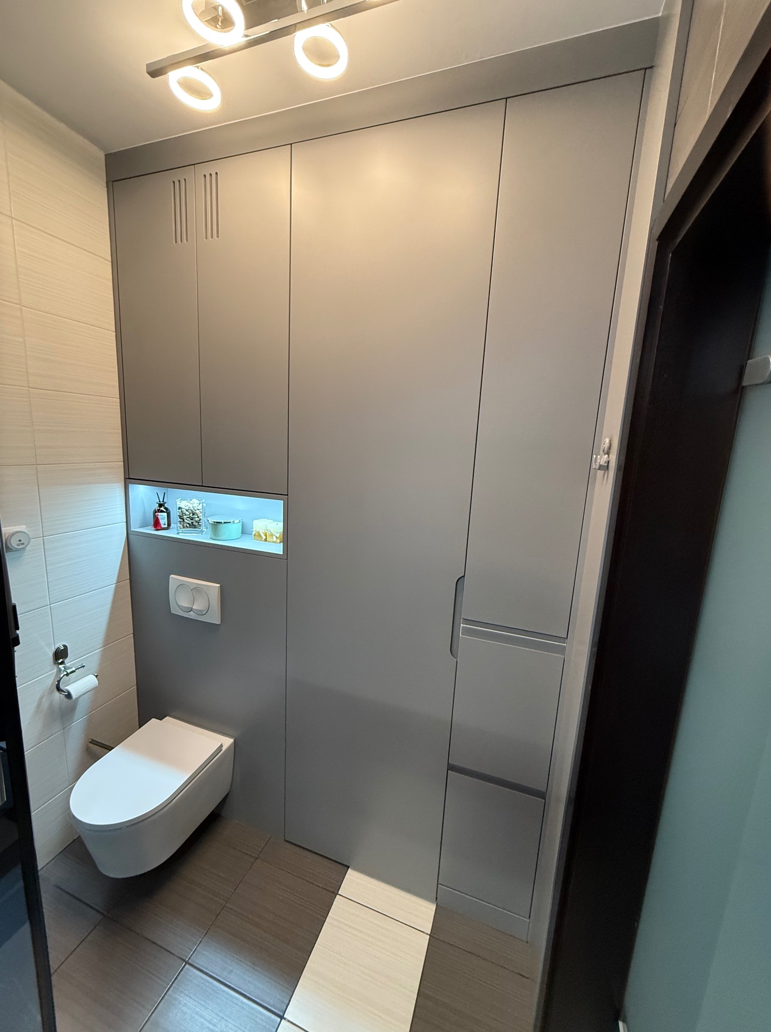 Szara szafa wnękowa w łazience z podświetlaną wnęką nad toaletą, dekoracje w wnęce.