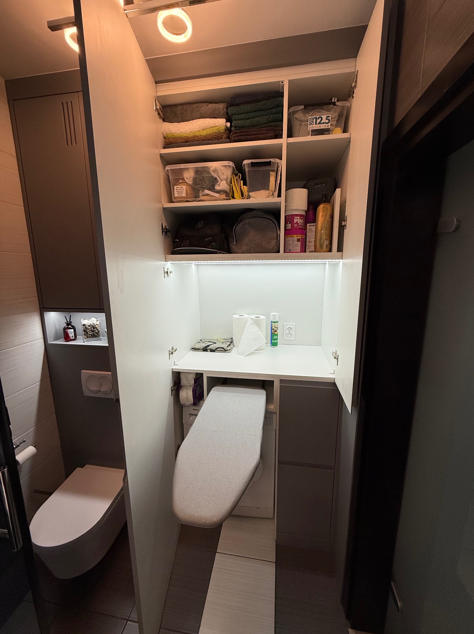 Wnętrze łazienki z zabudową na wymiar: szafki z półkami, pralka ukryta za frontem, deska do prasowania schowana w szafce, toaleta wisząca, oświetlenie LED.