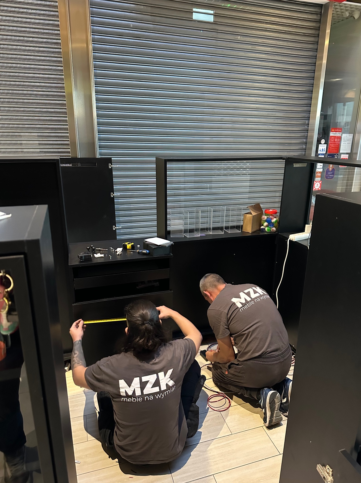 Dwóch monterów w szarych koszulkach z logo 'MZK meble na wymiar' mierzy czarne moduły meblowe w lokalu z zamkniętą metalową roletą.