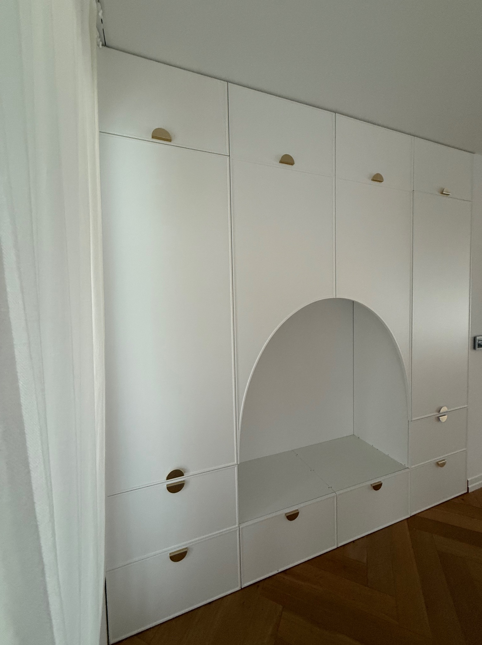Biała szafa na wymiar z półokrągłą wnęką, minimalistyczne złote uchwyty, parkiet w jodełkę na podłodze, delikatna zasłona w tle.