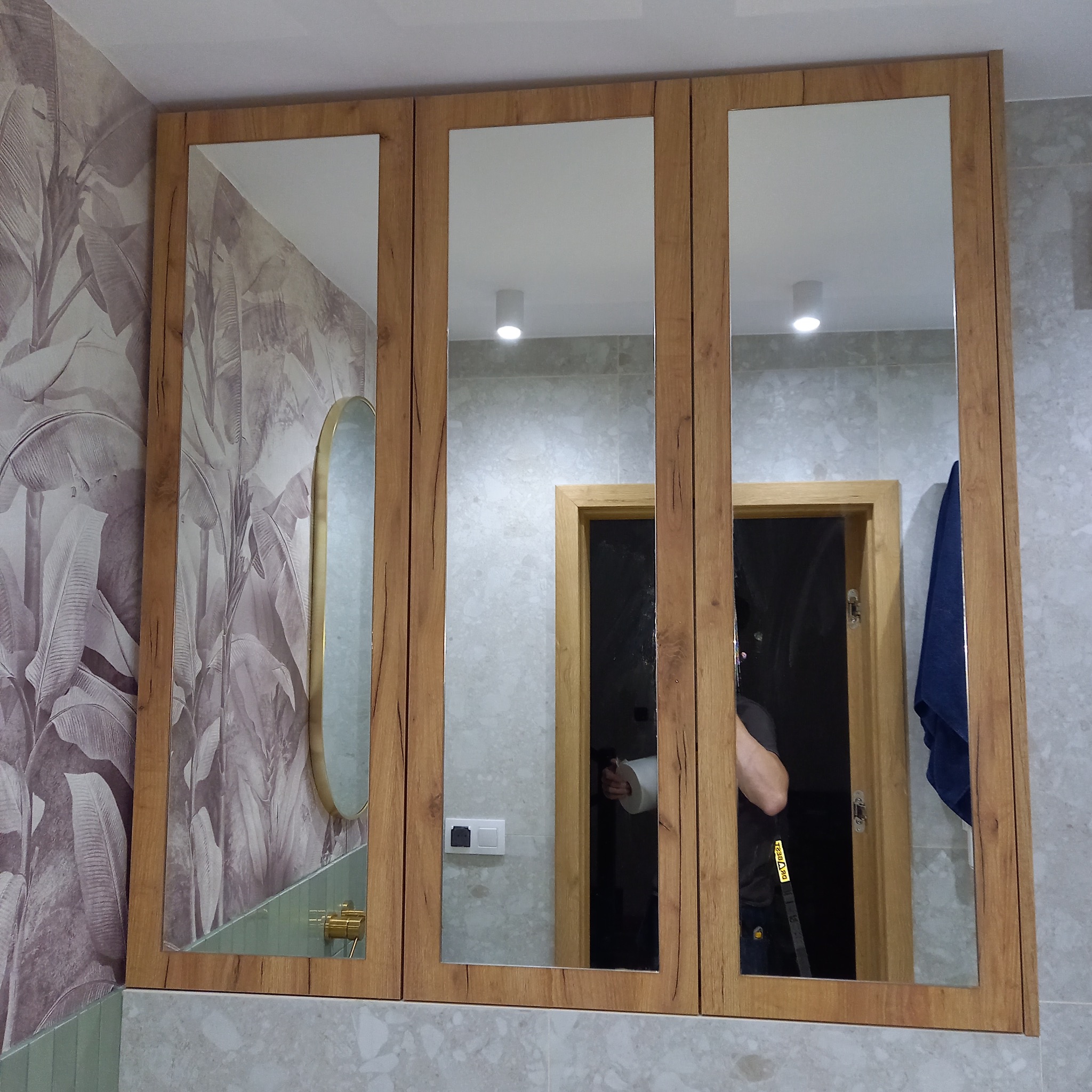 Trzyczęściowa szafka łazienkowa z lustrami i frontami w kolorze drewna, zamontowana nad umywalką z zielonymi płytkami, z widocznym fragmentem ściany w tapetę w liście i odbiciem osoby w lustrze.