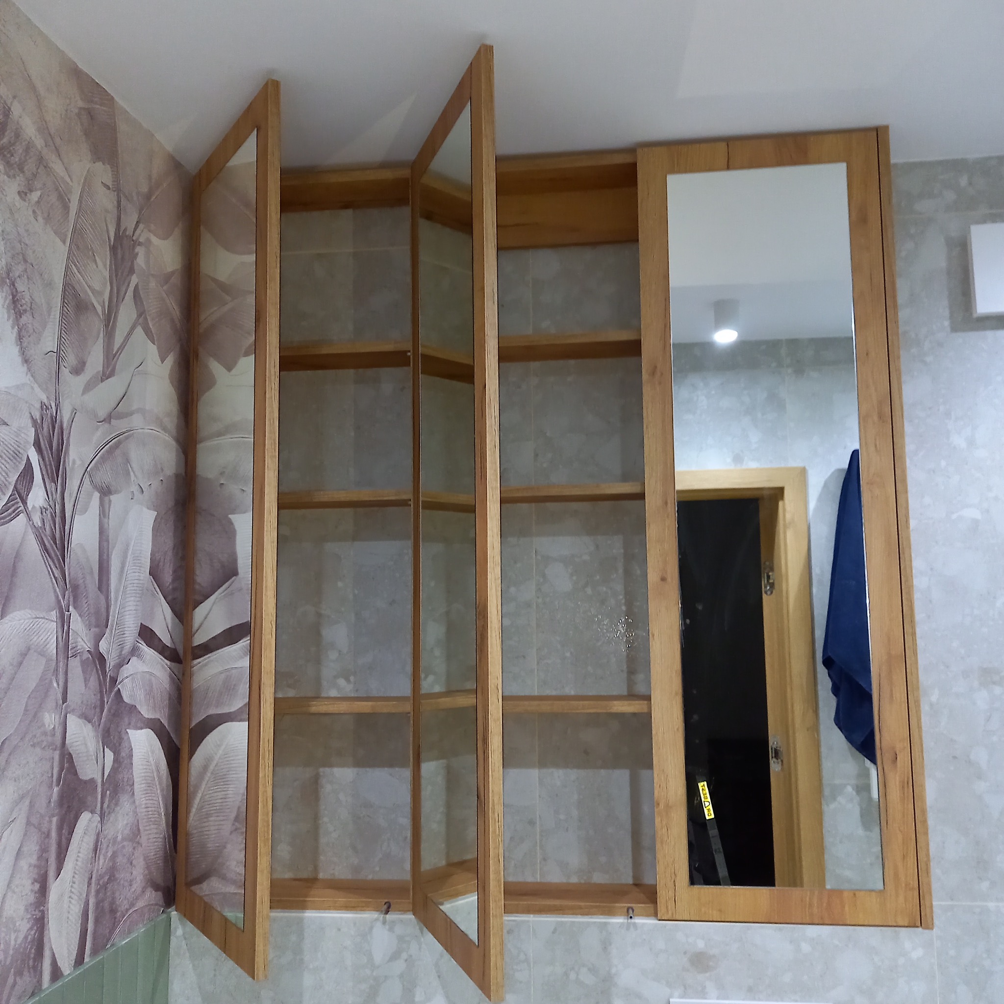 Drewniana szafka łazienkowa z lustrem, otwarte przeszklone drzwiczki, widoczna tapeta w liście i szara płytka na ścianie.