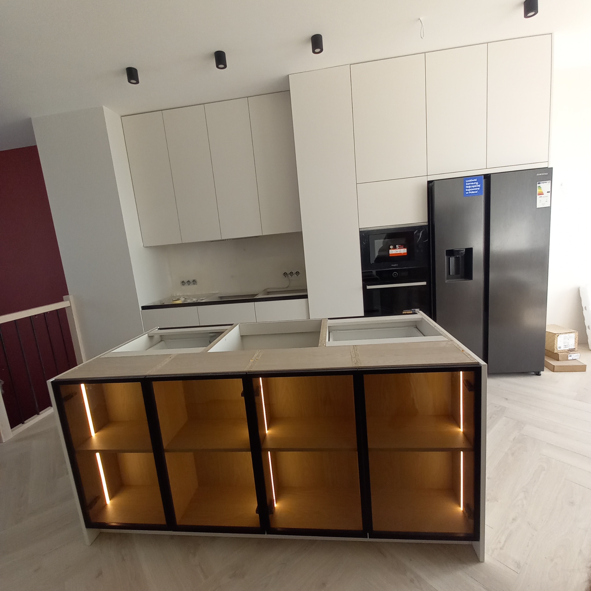 Wnętrze kuchni w trakcie montażu z wyspą z podświetlanymi półkami, widoczne białe szafki wiszące i sprzęt AGD.
