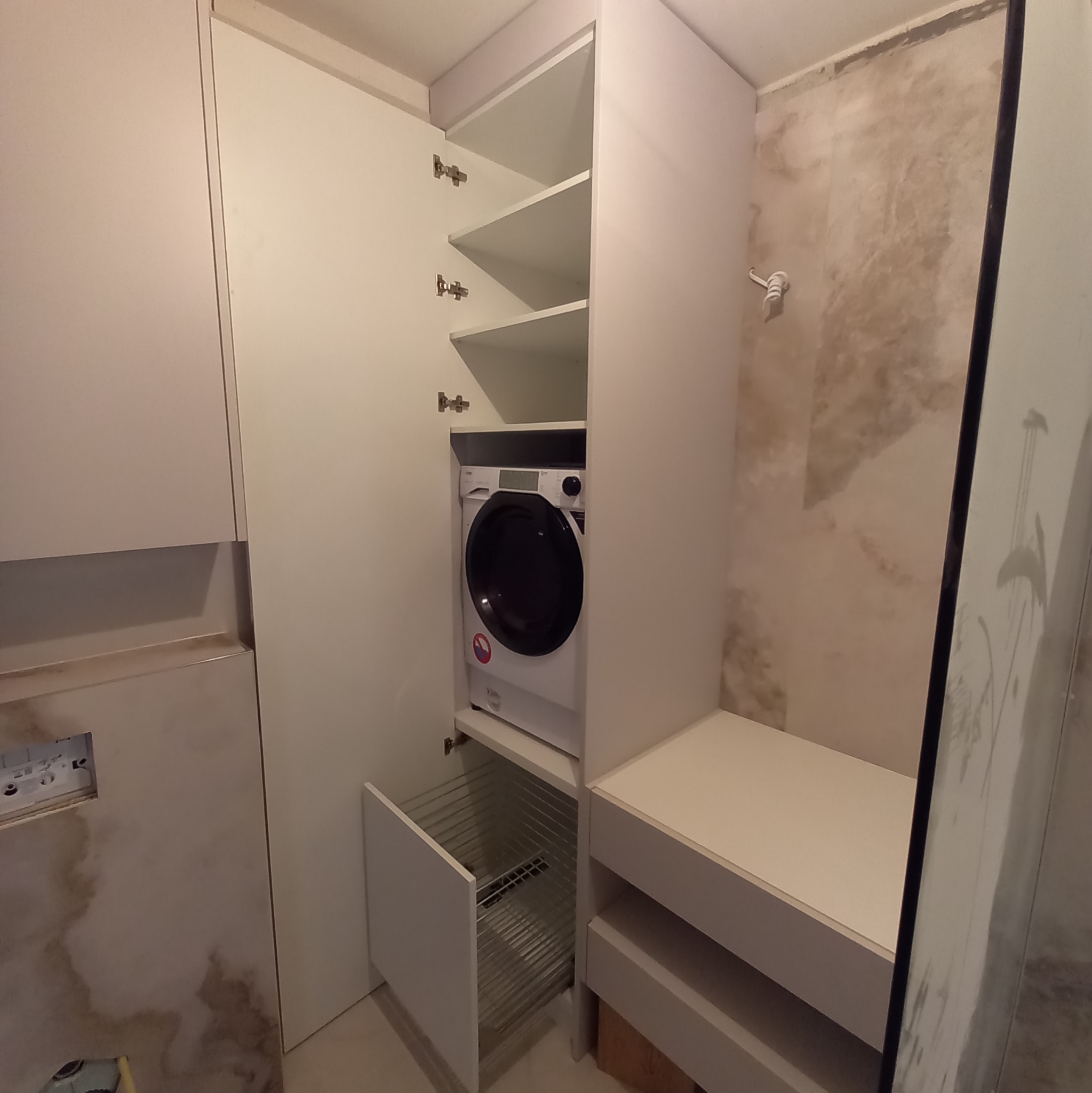 Biała zabudowa meblowa z pralką w szafie i wysuwanym koszem na pranie, widoczne półki i fragment ściany z surowym wykończeniem.