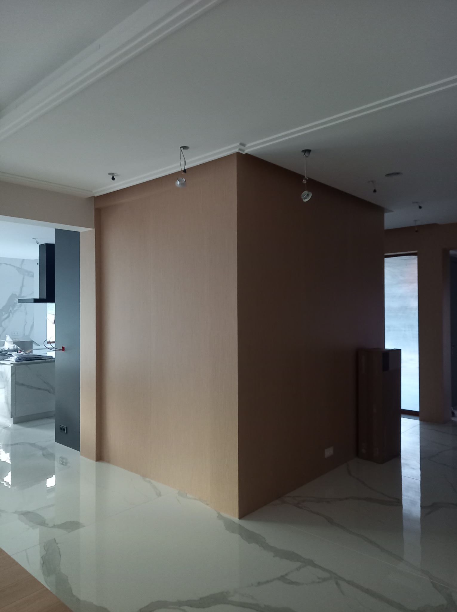 Wnętrze w trakcie wykańczania: widoczna ściana w kolorze beżowym, błyszcząca podłoga z marmurowym wzorem, fragment kuchni z szarymi szafkami i białym blatem, oraz kartony stojące przy oknie.