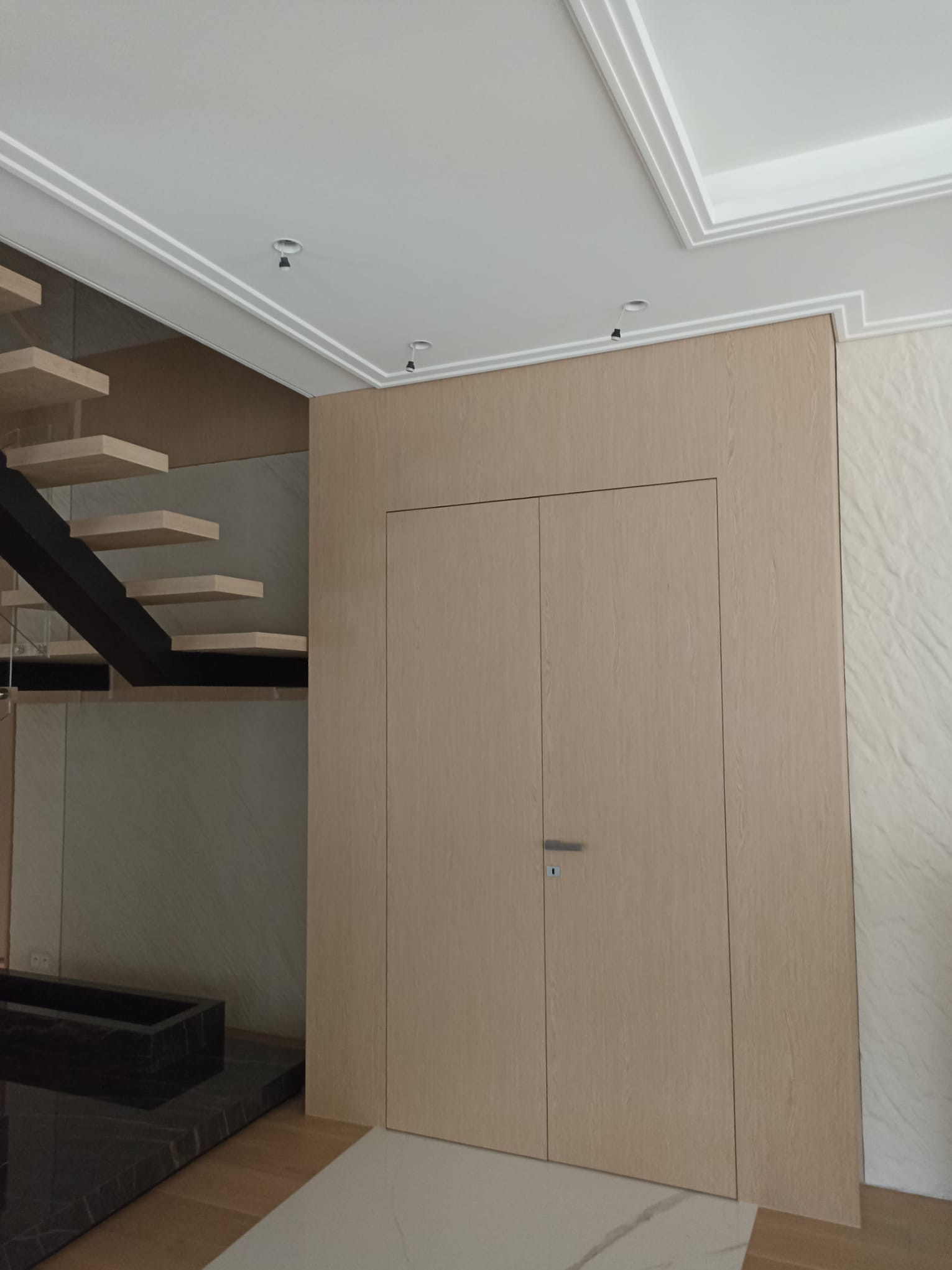 Ukryte drzwi w zabudowie meblowej z jasnego drewna, zintegrowane ze ścianą, obok nowoczesnych schodów z drewnianymi stopniami i czarną konstrukcją, widoczne oświetlenie punktowe na suficie.
