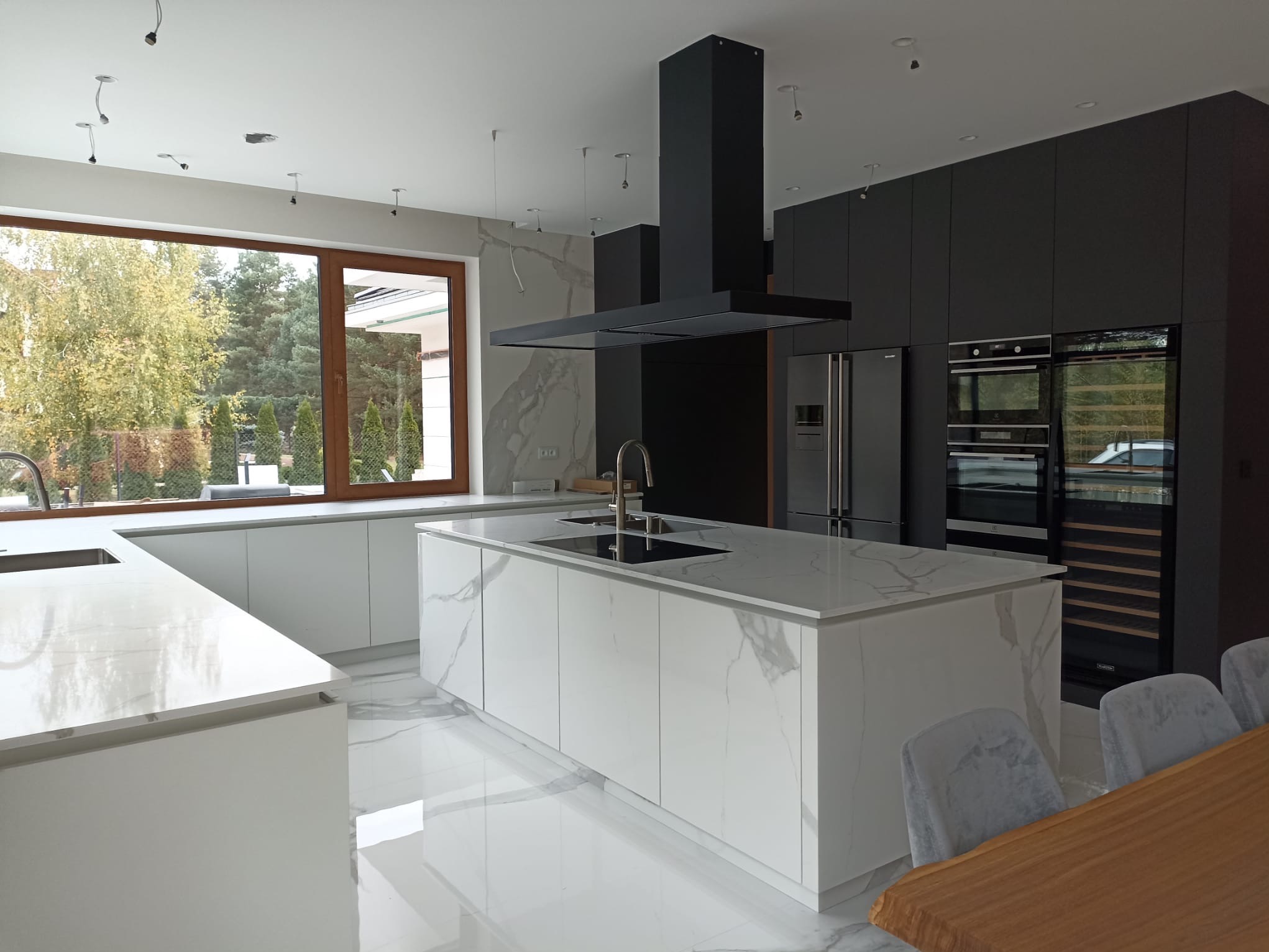 Nowoczesna, minimalistyczna kuchnia z wyspą wykończoną białym marmurem, czarnym okapem i zabudową w odcieniach szarości; widoczne duże okno z widokiem na zieleń.
