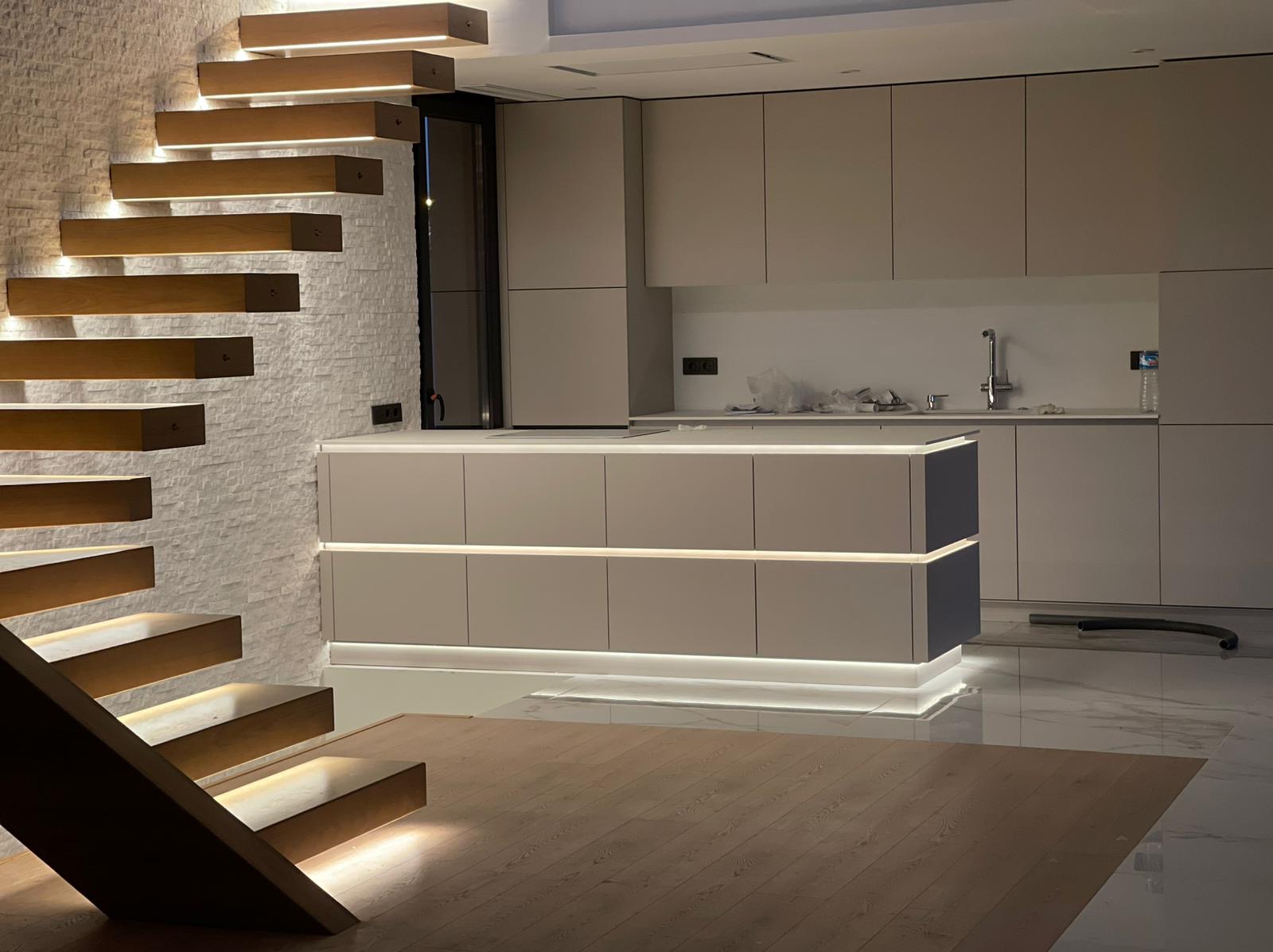 Nowoczesna kuchnia z wyspą i podświetleniem LED, drewniane schody z oświetleniem punktowym, biała ściana z cegły, parkiet w jodełkę, marmurowe płytki na podłodze.