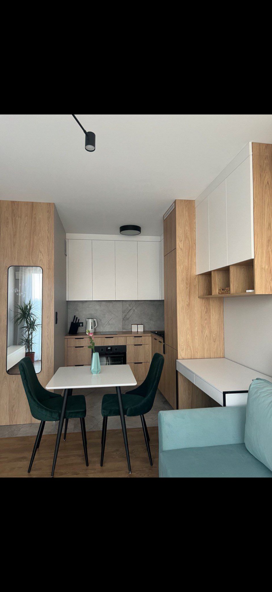 Nowoczesna kuchnia z drewnianymi i białymi frontami, szarym blatem i marmurowym backsplashiem, uzupełniona o stół z dwoma zielonymi krzesłami i biurko z półkami, całość w jasnym, minimalistycznym...