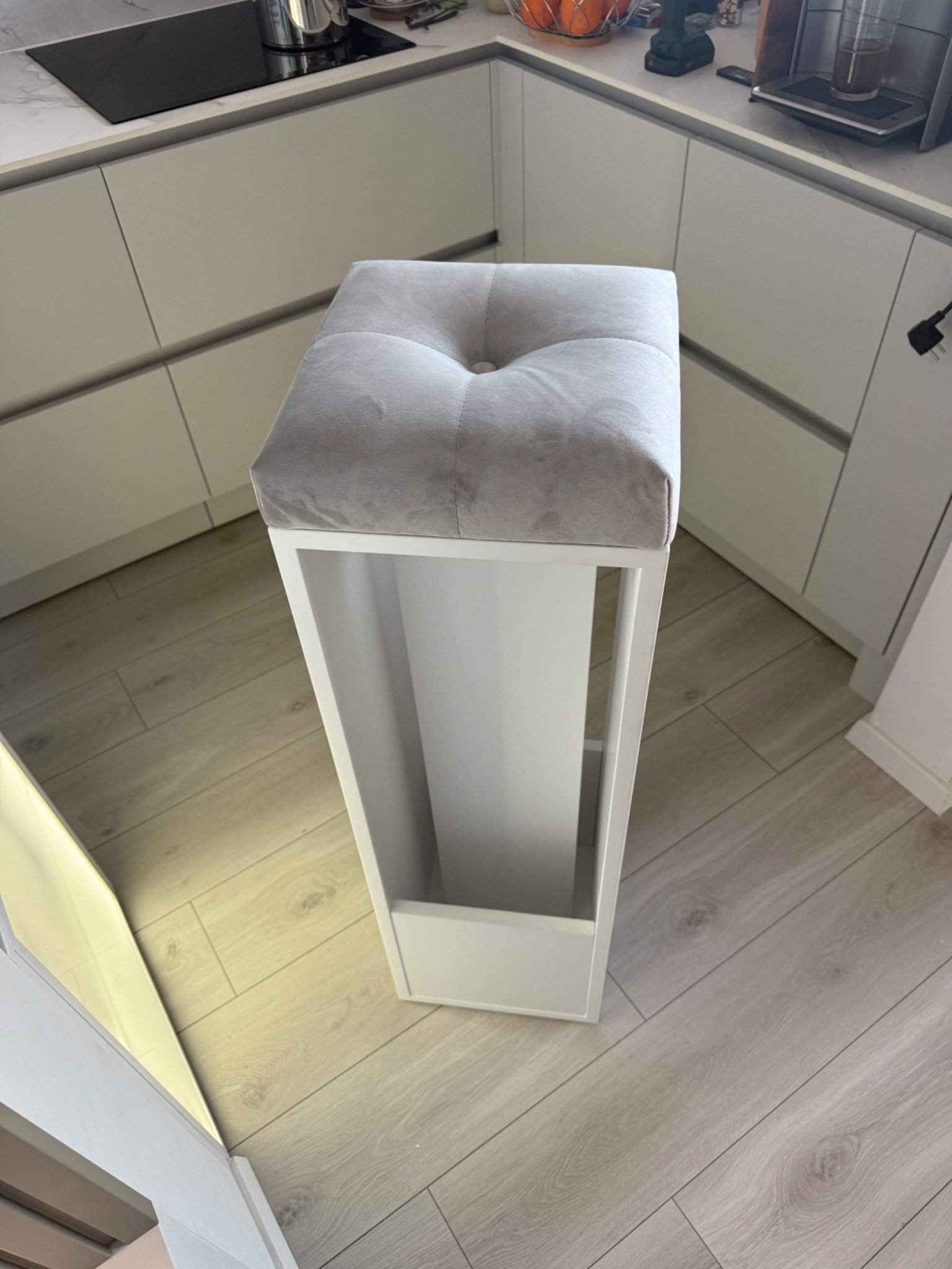 Biały stołek barowy z szarą, tapicerowaną poduszką siedziska, ustawiony w nowoczesnej kuchni z białymi frontami szafek i jasną podłogą z paneli.