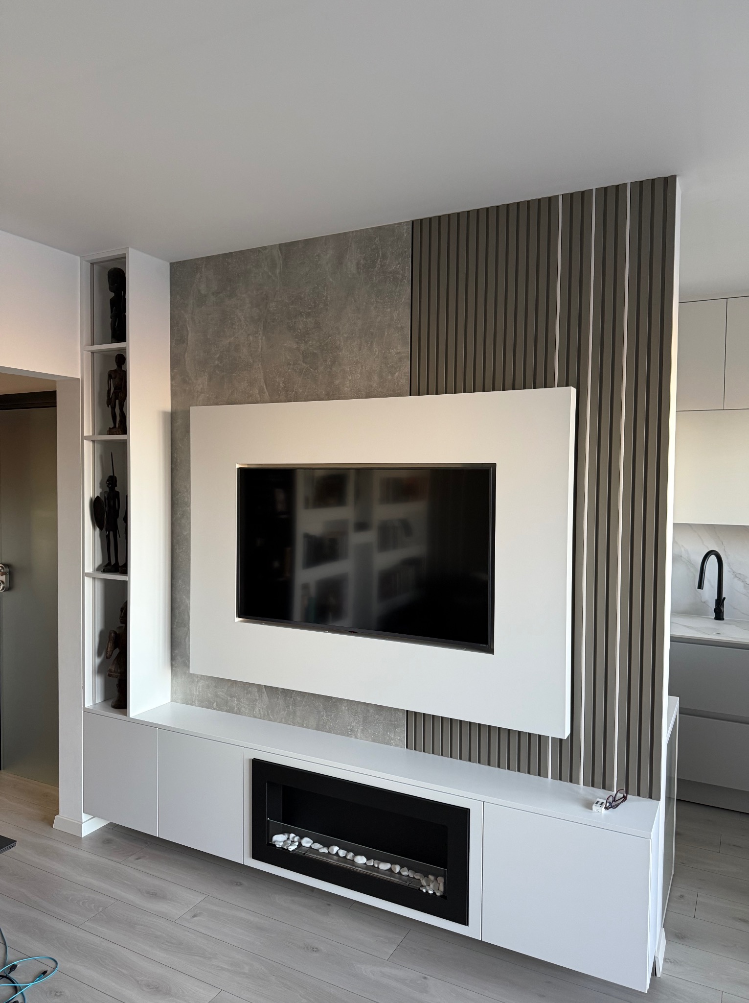 Nowoczesna zabudowa ściany z telewizorem i kominkiem elektrycznym, białe szafki, dekoracyjne panele ścienne i półki z afrykańskimi rzeźbami, widok na kuchnię w tle.