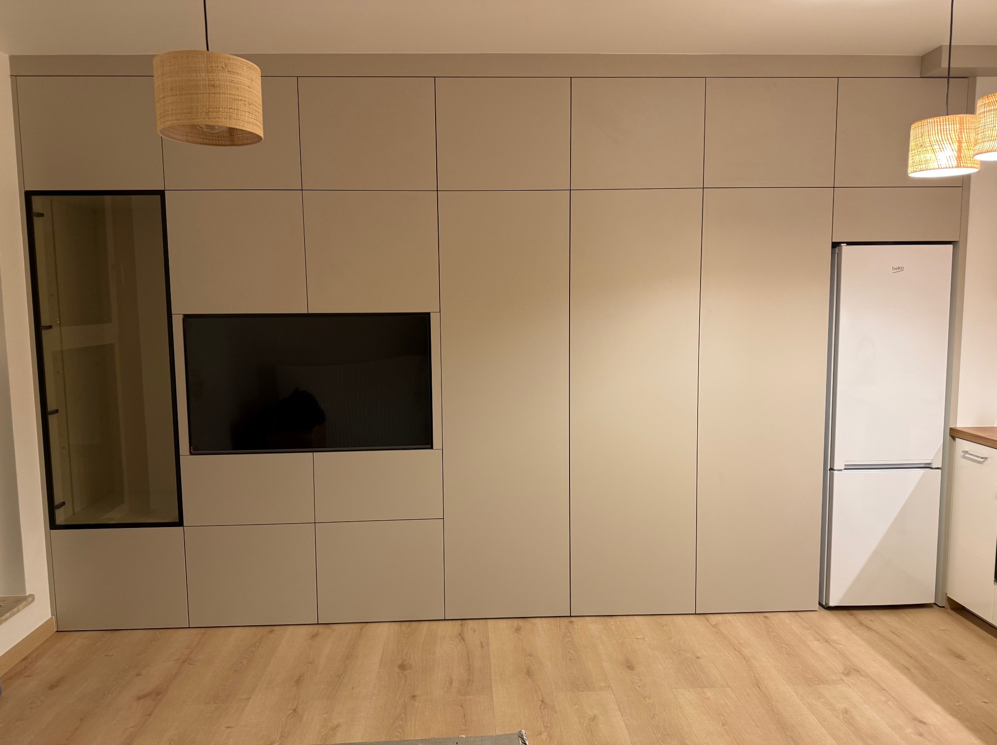 Beżowa zabudowa meblowa w salonie z wbudowanym telewizorem, przeszkloną witryną i lodówką, oświetlona wiszącymi lampami z abażurami z plecionki.