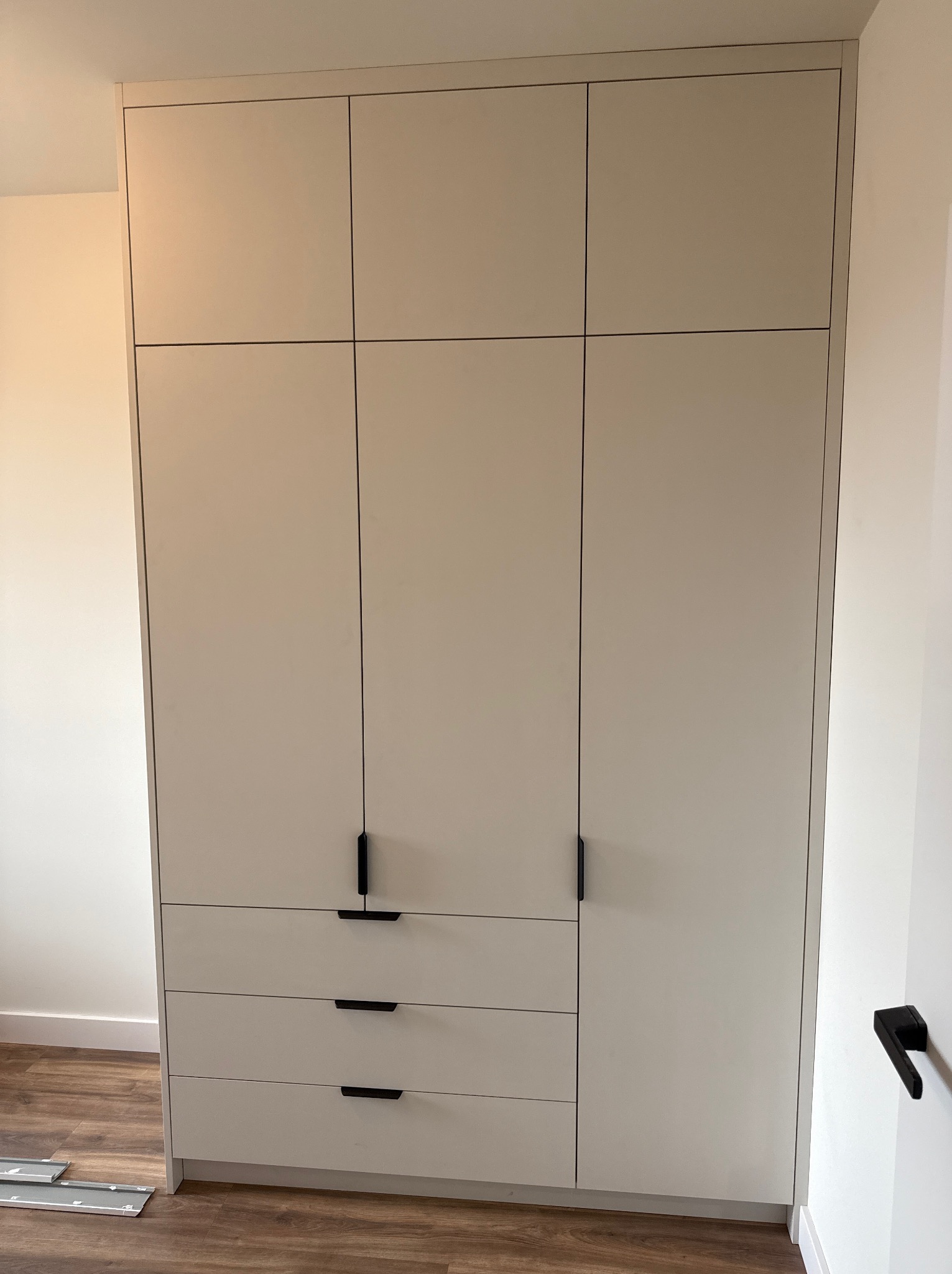 Wysoka, zabudowana szafa z białymi frontami, czarnymi, minimalistycznymi uchwytami, z trzema szufladami na dole i szafkami zamykanymi na górze, widok na fragment drewnianej podłogi.