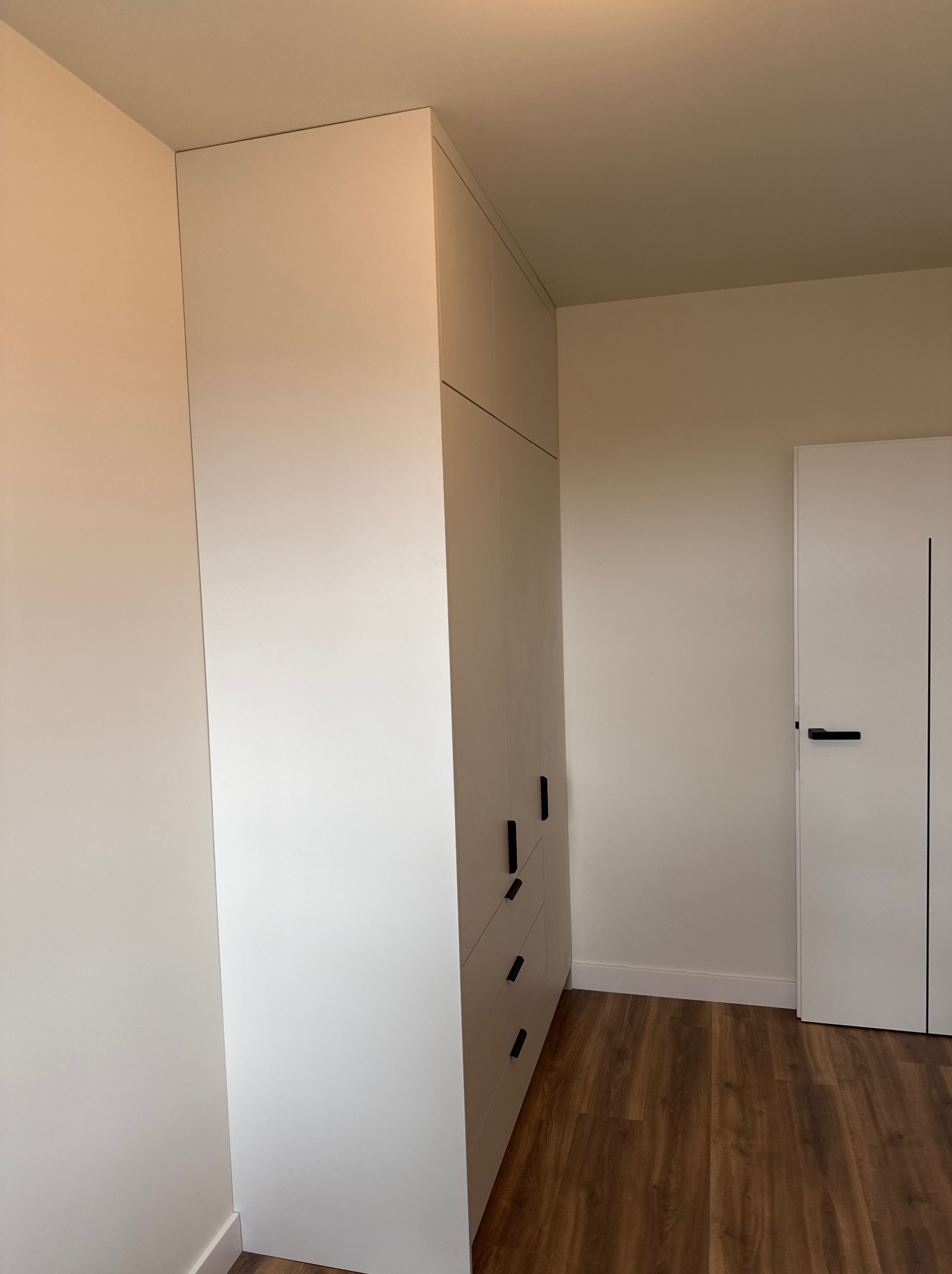 Wysoka, biała szafa wnękowa z czarnymi, minimalistycznymi uchwytami, dopasowana do sufitu, na tle białej ściany i drewnianej podłogi, obok białe drzwi z czarnym akcentem.