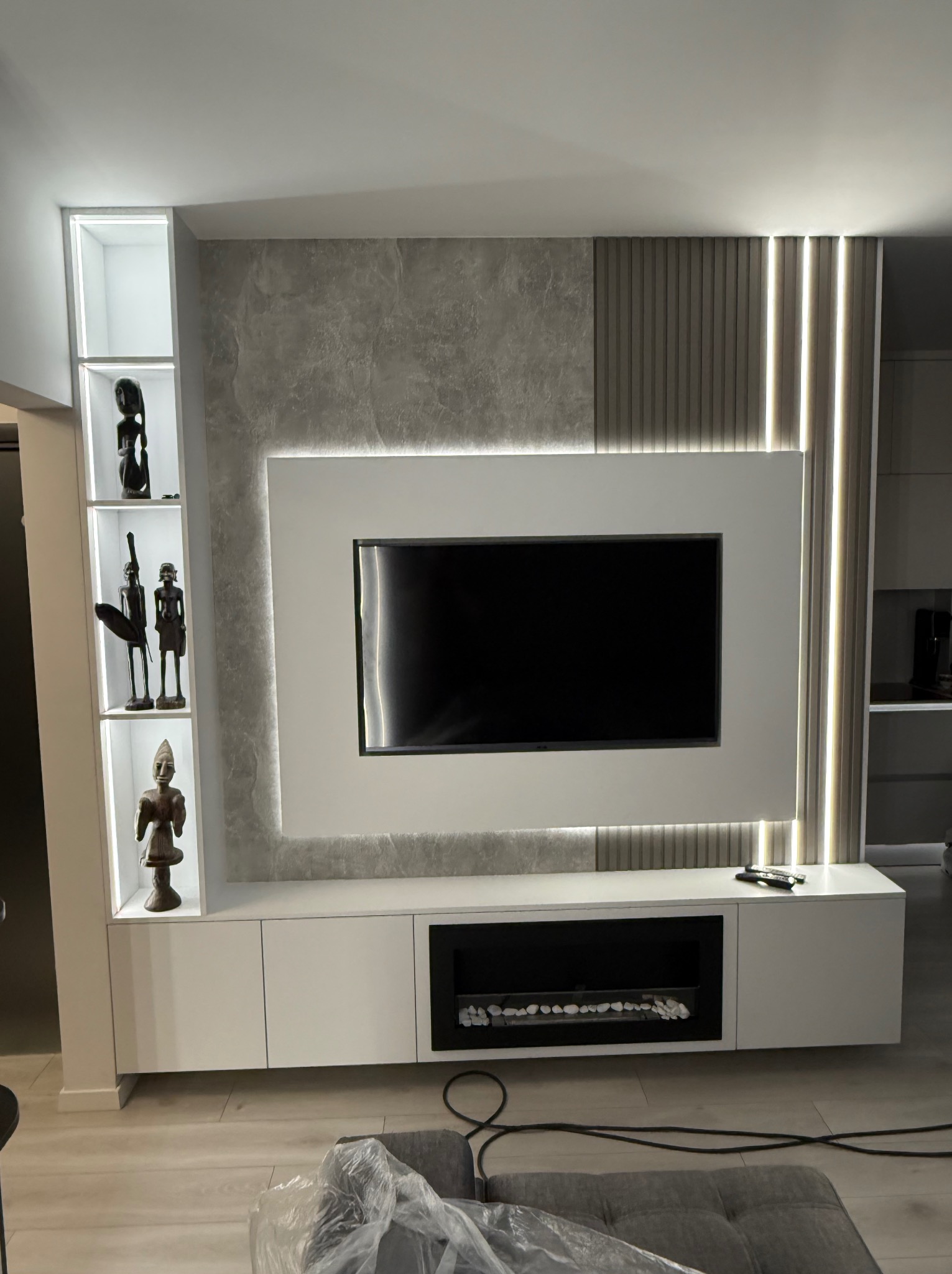 Nowoczesna ściana telewizyjna z podświetleniem LED, dekoracyjnym panelem imitującym marmur, pionowymi lamelami i minimalistyczną szafką RTV z kominkiem elektrycznym oraz półkami z afrykańskimi...