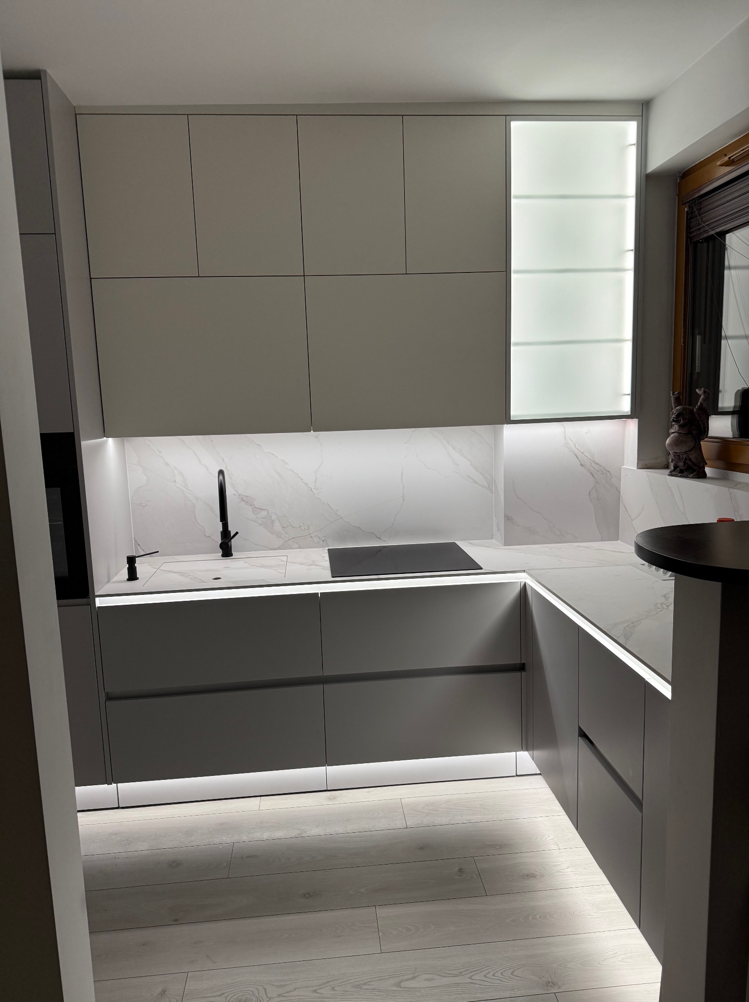 Nowoczesna, szara kuchnia z białym blatem imitującym marmur, podświetleniem LED i czarną armaturą, z widoczną figurką Buddy na parapecie okna.