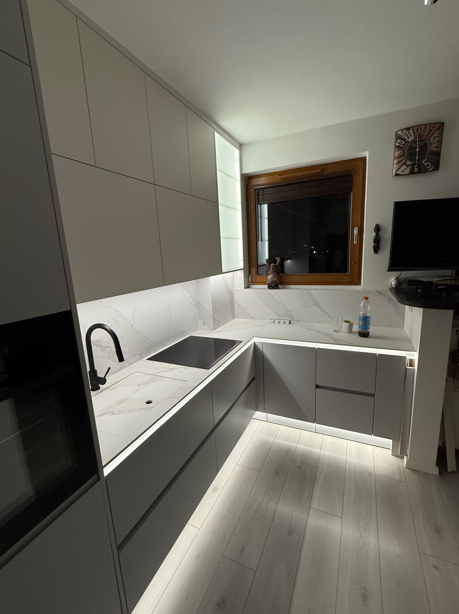 Nowoczesna kuchnia w odcieniach szarości z marmurowym blatem i podświetleniem LED pod szafkami, widok na okno z nocnym krajobrazem.