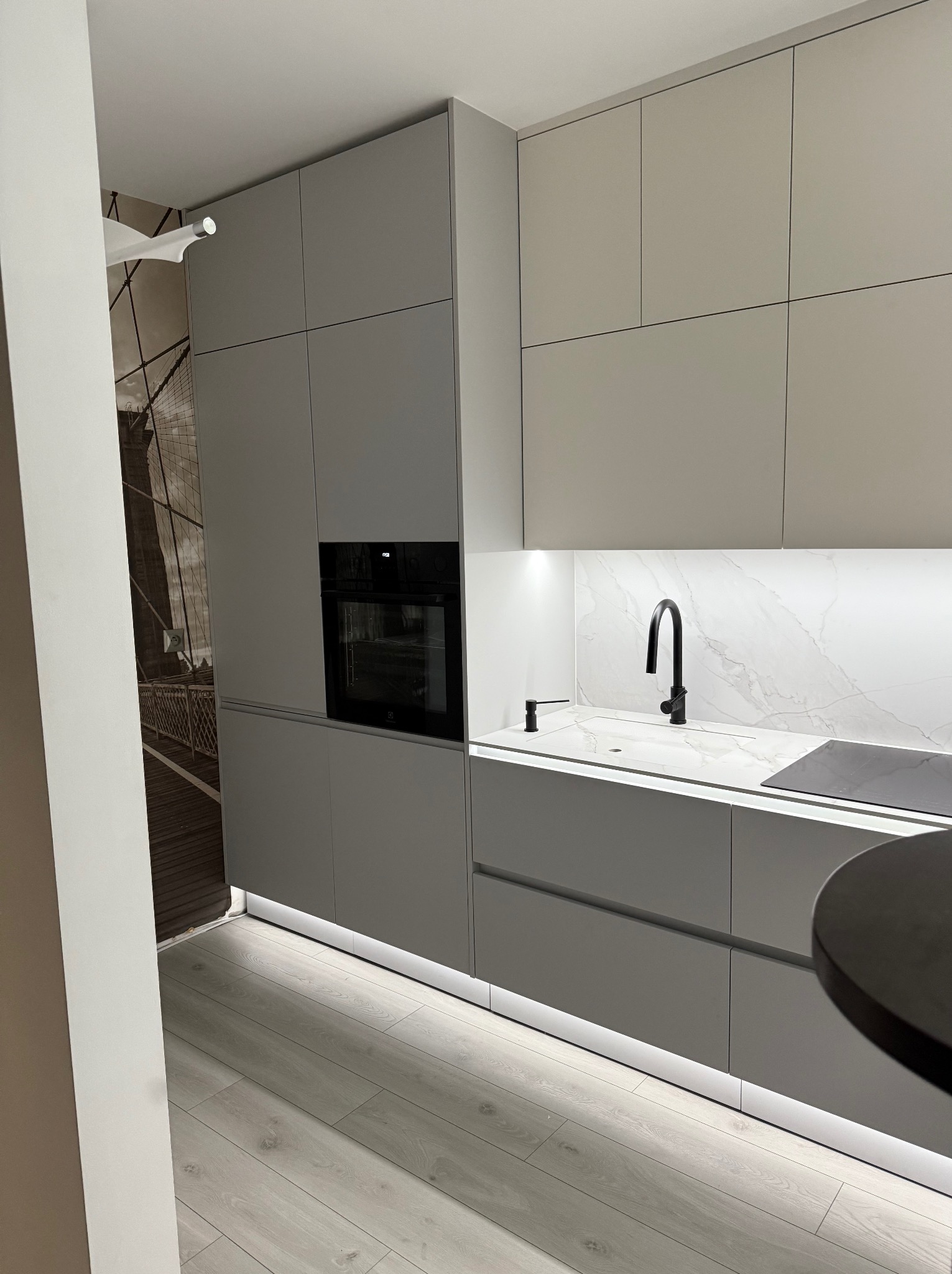 Nowoczesna, szara kuchnia z podświetleniem LED pod szafkami, czarną armaturą i piekarnikiem w zabudowie, z tapetą przedstawiającą most w tle.