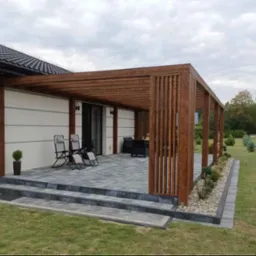 Nowoczesna pergola z ciemnego drewna BSH, z pionowymi listwami z jednej strony, zadaszająca taras wyłożony szarą kostką, z dwoma leżakami i zestawem mebli ogrodowych w tle, na tle białej ściany...