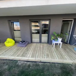 Taras drewniany na parterze mieszkania z jasnego drewna, z widoczną pufą w kolorze neonowej zieleni, stolikiem z kwiatami doniczkowymi i suszarką na pranie.