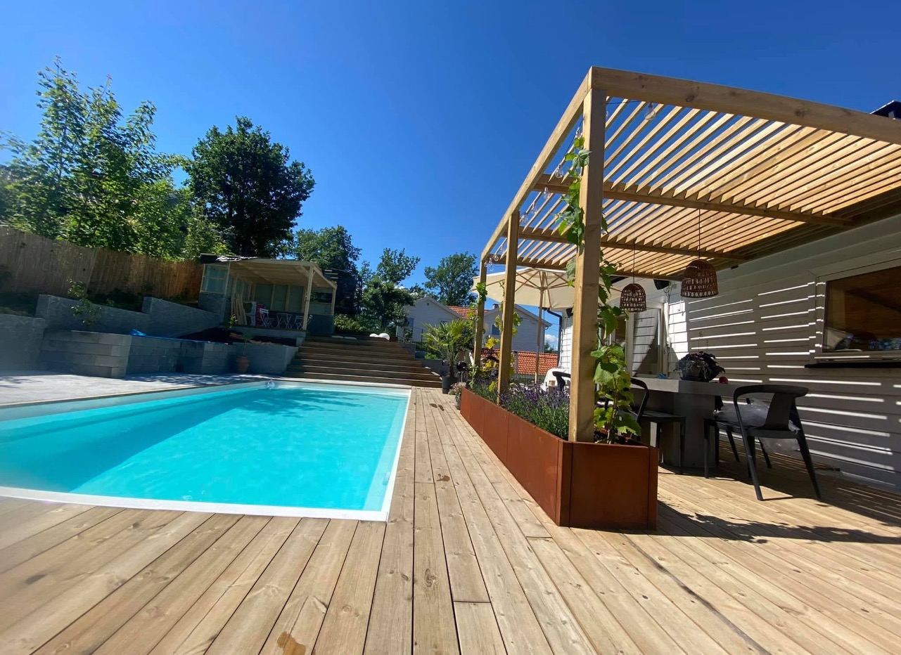 Drewniany taras z basenem, pergola z pnączami, rustykalna donica z lawendą, widok na altanę w tle.