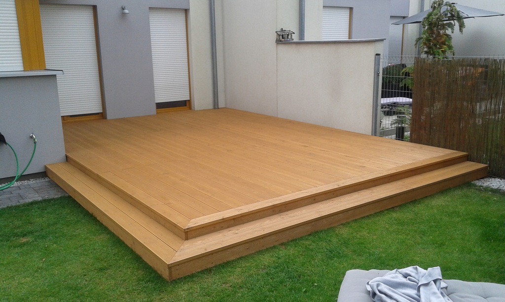 Nowoczesny taras z drewna z dwoma stopniami, przylegający do jasnego budynku z roletami zewnętrznymi, otoczony zielonym trawnikiem.