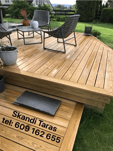 Drewniany taras z meblami ogrodowymi i roślinami w donicach, widoczny fragment schodów z ciemną płytką i numerem telefonu.