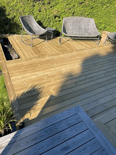 Jasna drewniana podłoga tarasu z szarymi, plecionymi krzesłami i stolikiem kawowym na tle zielonego żywopłotu; widoczny fragment niebieskoszarego tarasu na pierwszym planie.