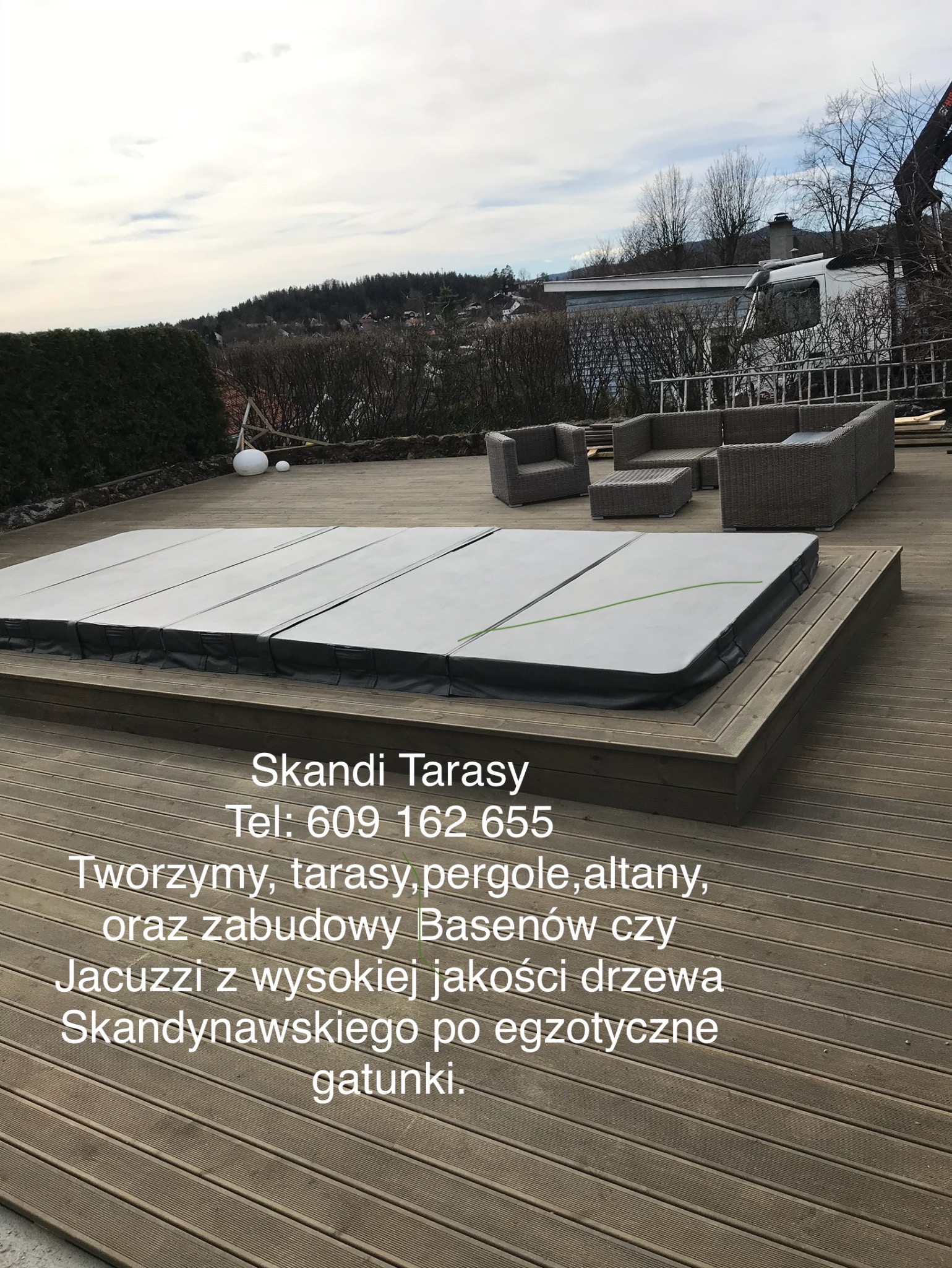 Zadaszone jacuzzi wbudowane w drewniany taras, otoczone meblami ogrodowymi z rattanu, w tle widok na zieleń i zabudowania.