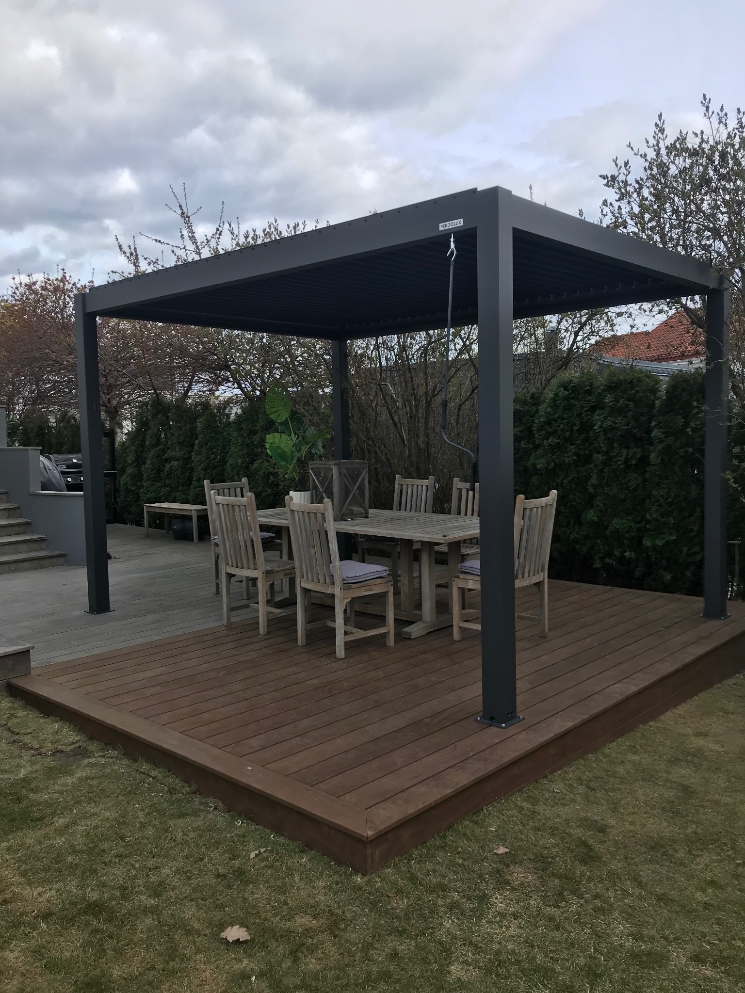 Nowoczesna, grafitowa pergola z aluminiową konstrukcją na tarasie z desek w ciepłym odcieniu brązu; na tarasie stół z krzesłami, w tle zielone tuje.