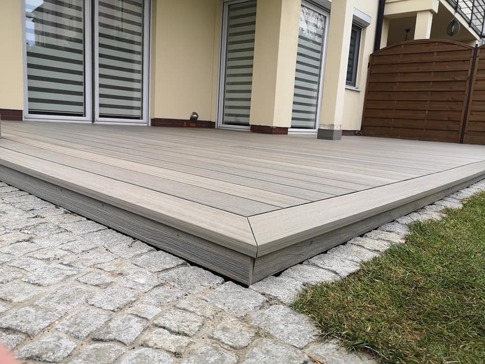 Jasnoszary taras kompozytowy z widoczną strukturą drewna, obrzeże wykończone kostką granitową i fragmentem trawnika, w tle okna z żaluzjami poziomymi i fragment drewnianego ogrodzenia.