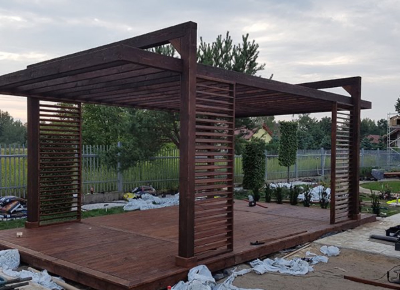 Ciemnobrązowa drewniana pergola z ażurowymi ścianami na drewnianym tarasie w trakcie budowy, widoczne narzędzia i worki z materiałami.