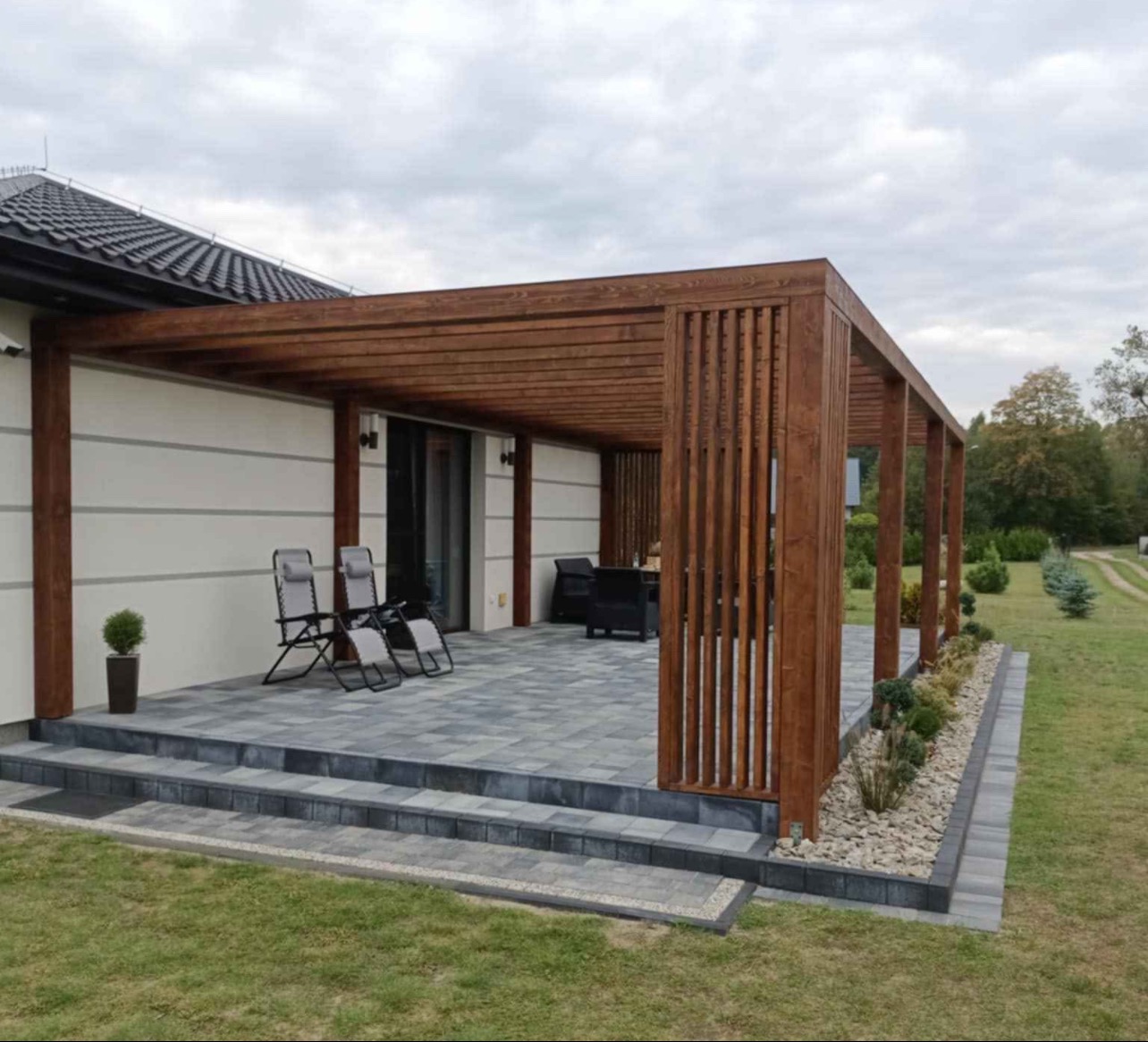 Nowoczesna pergola z ciemnego drewna BSH, z pionowymi listwami z jednej strony, zadaszająca taras wyłożony szarą kostką, z dwoma leżakami i zestawem mebli ogrodowych w tle, na tle białej ściany...