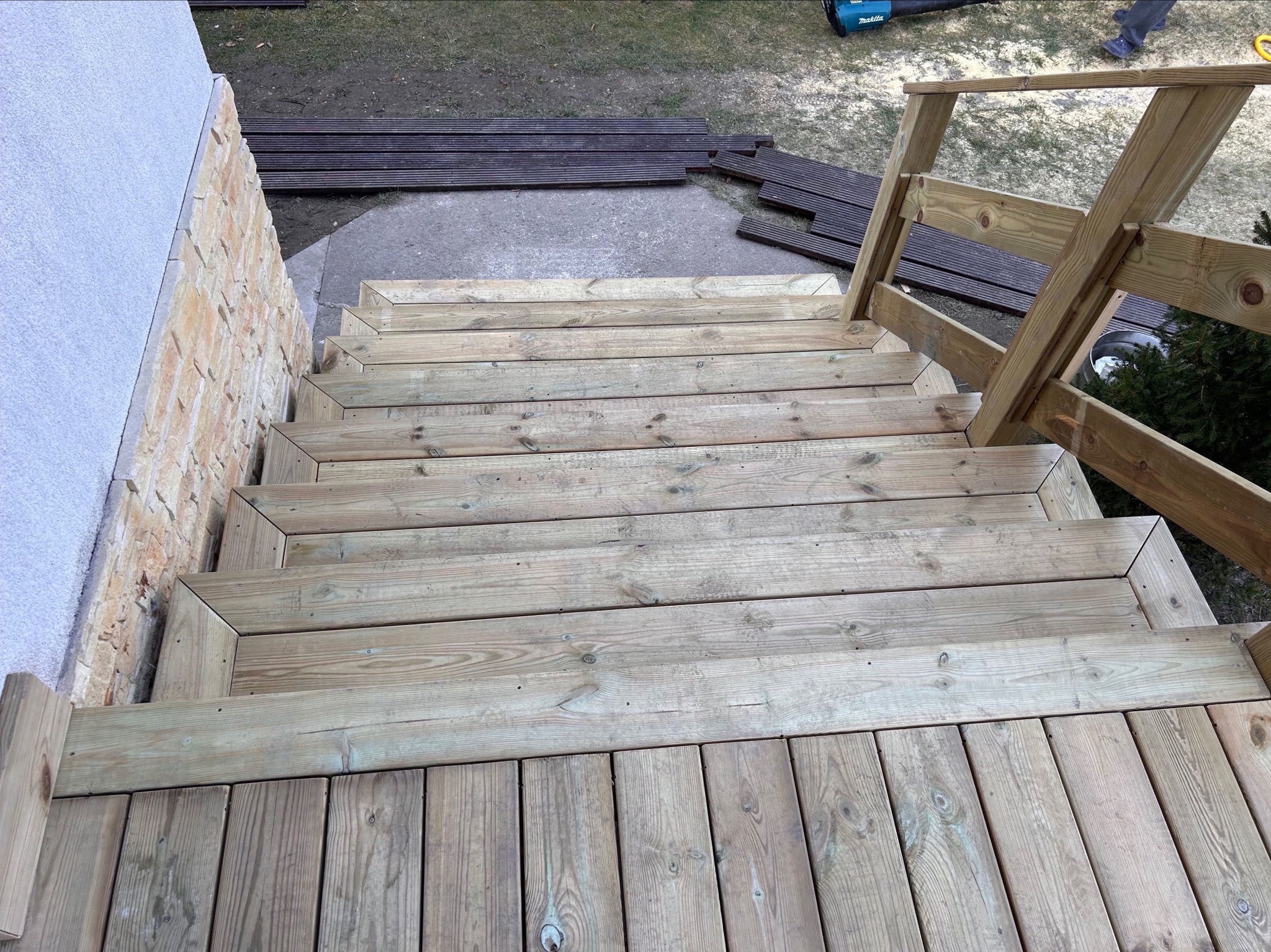 Drewniane schody prowadzące z tarasu, z jasnego drewna, z prostą balustradą, widoczne deski tarasowe i narzędzie budowlane w tle.