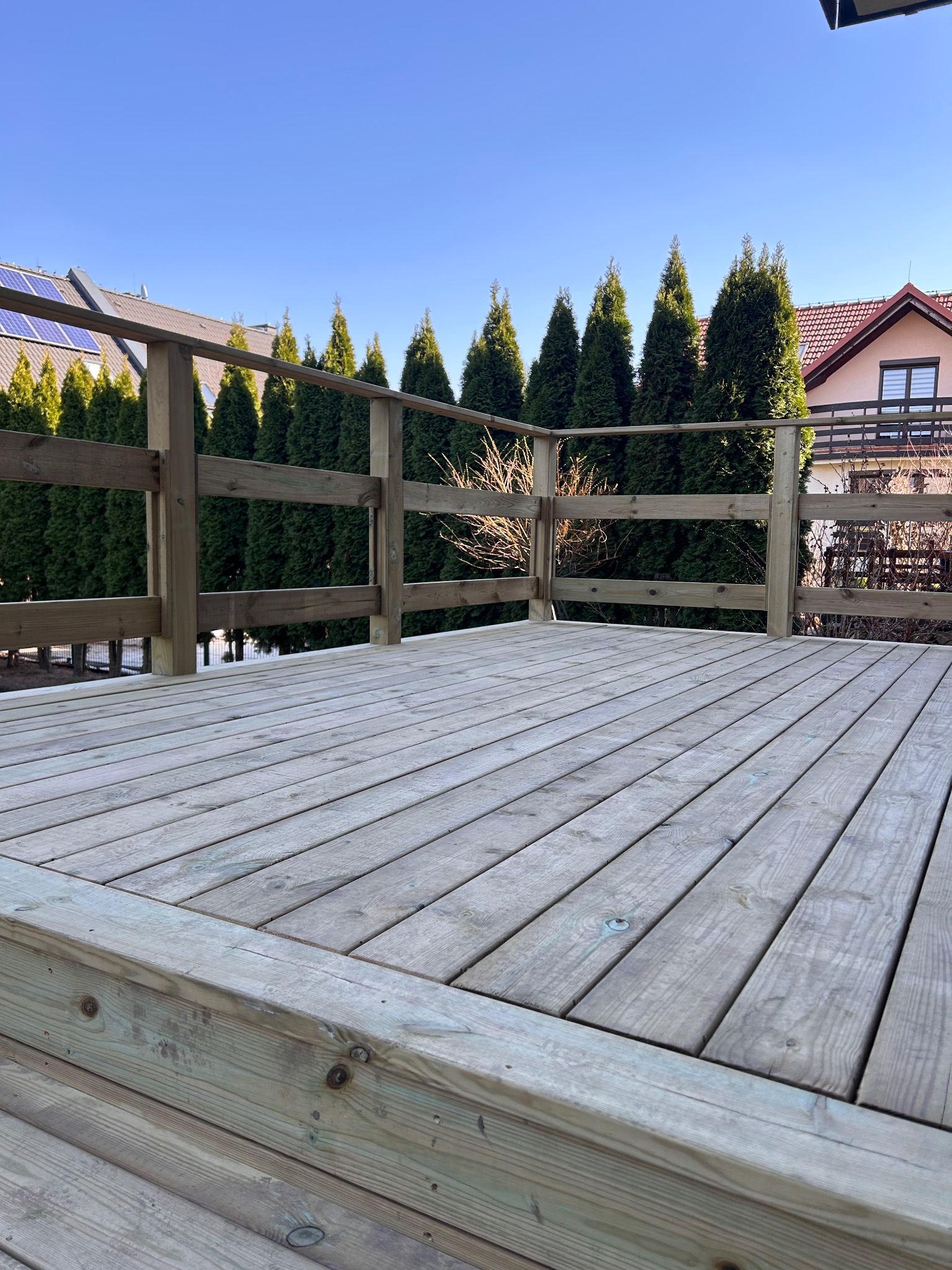 Nowy drewniany taras z jasnych desek z drewnianą balustradą, w tle rząd wysokich, zielonych tuj i dom z czerwonym dachem w słoneczny dzień.