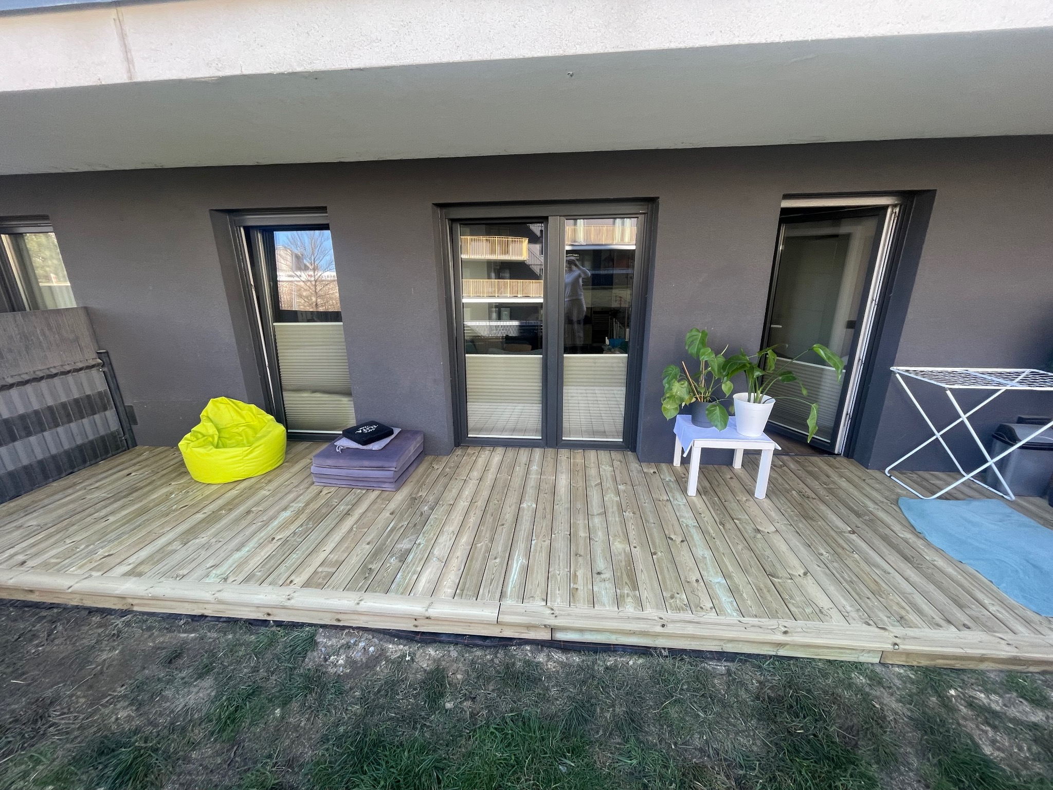 Taras drewniany na parterze mieszkania z jasnego drewna, z widoczną pufą w kolorze neonowej zieleni, stolikiem z kwiatami doniczkowymi i suszarką na pranie.