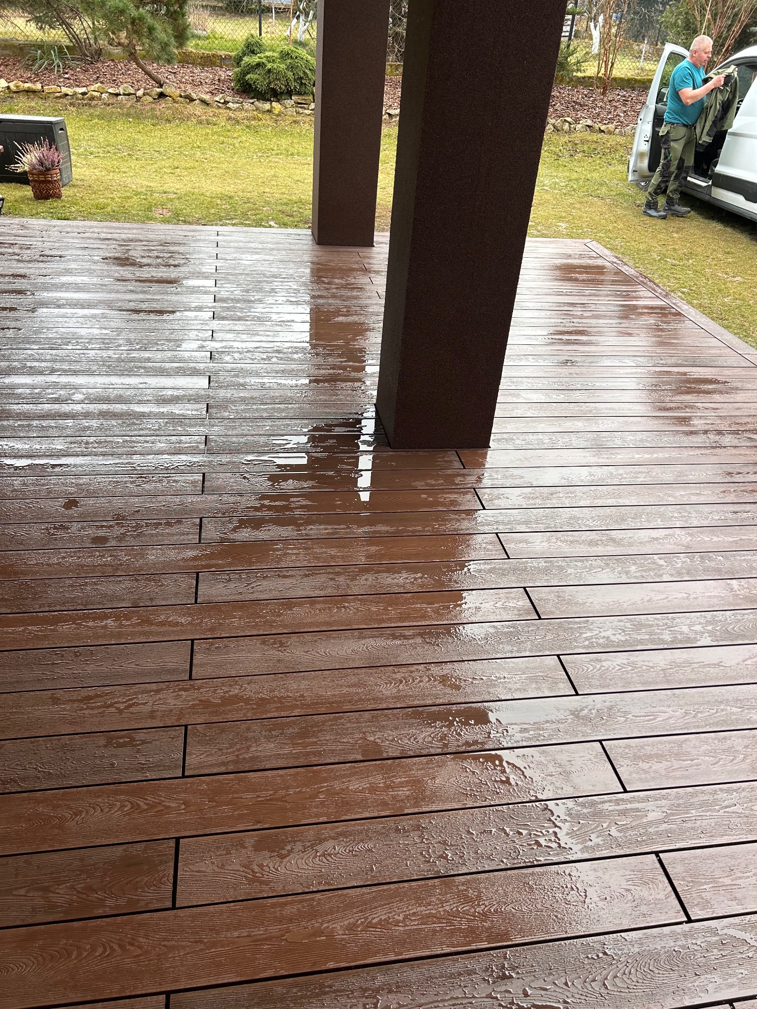 Mokry, brązowy taras z desek kompozytowych po deszczu, z widocznymi kroplami wody i odbiciami, z fragmentem filaru i pracownikiem wyjmującym coś z samochodu w tle.