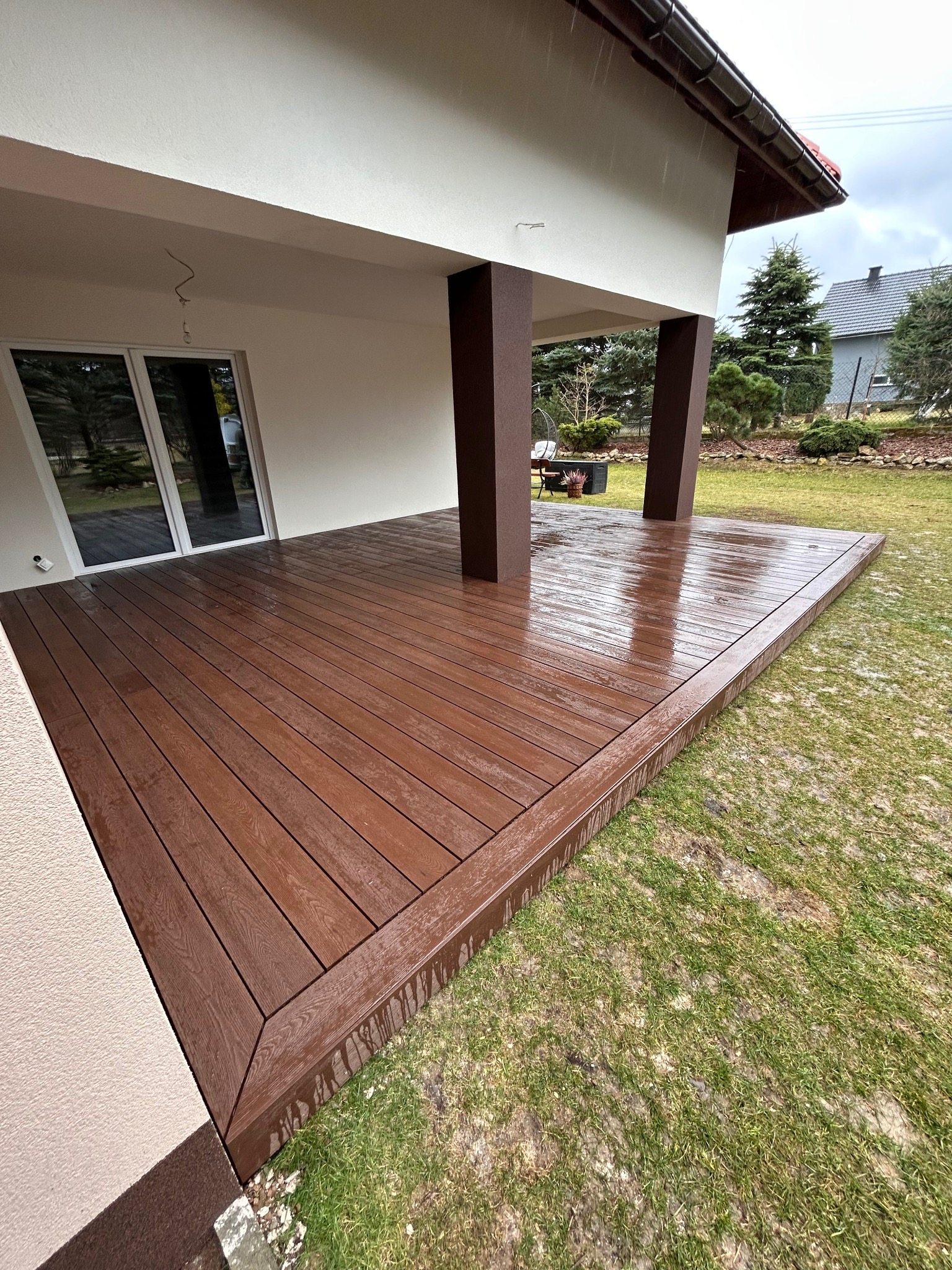 Zadaszony taras z deski kompozytowej w kolorze ciemnego drewna, wykończony obwodową deską, widok na ogród w pochmurny dzień.