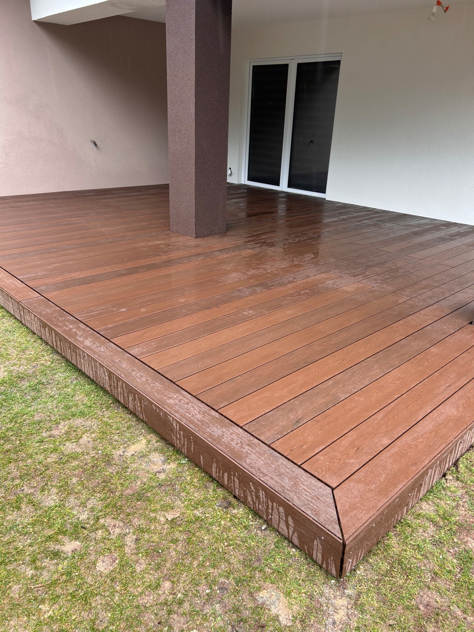 Nowoczesny taras z deski kompozytowej w odcieniach brązu, z zabudową boczną, widok na trawnik i ścianę budynku z oknem.