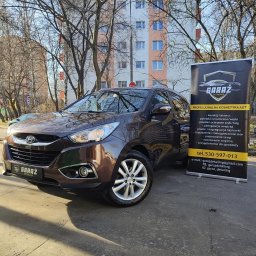 Garaz Auto Detailing Lublin - Brązowy SUV Hyundai zaparkowany na zewnątrz, obok niego baner reklamowy firmy detailingowej z ofertą korekty lakieru, powłok ochronnych, pielęgnacji wnętrza, prania tapicerki i ozonowania.