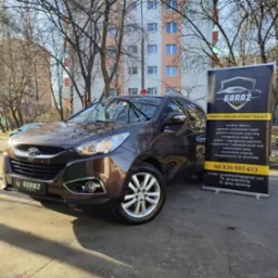 Brązowy SUV Hyundai zaparkowany na zewnątrz, obok niego baner reklamowy firmy detailingowej z ofertą korekty lakieru, powłok ochronnych, pielęgnacji wnętrza, prania tapicerki i ozonowania.