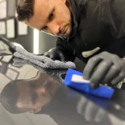 Detailer w czarnych rękawiczkach poleruje lakier ciemnego samochodu przy użyciu niebieskiego aplikatora i szarej mikrofibry, widoczne odbicie twarzy detailera w lakierze.